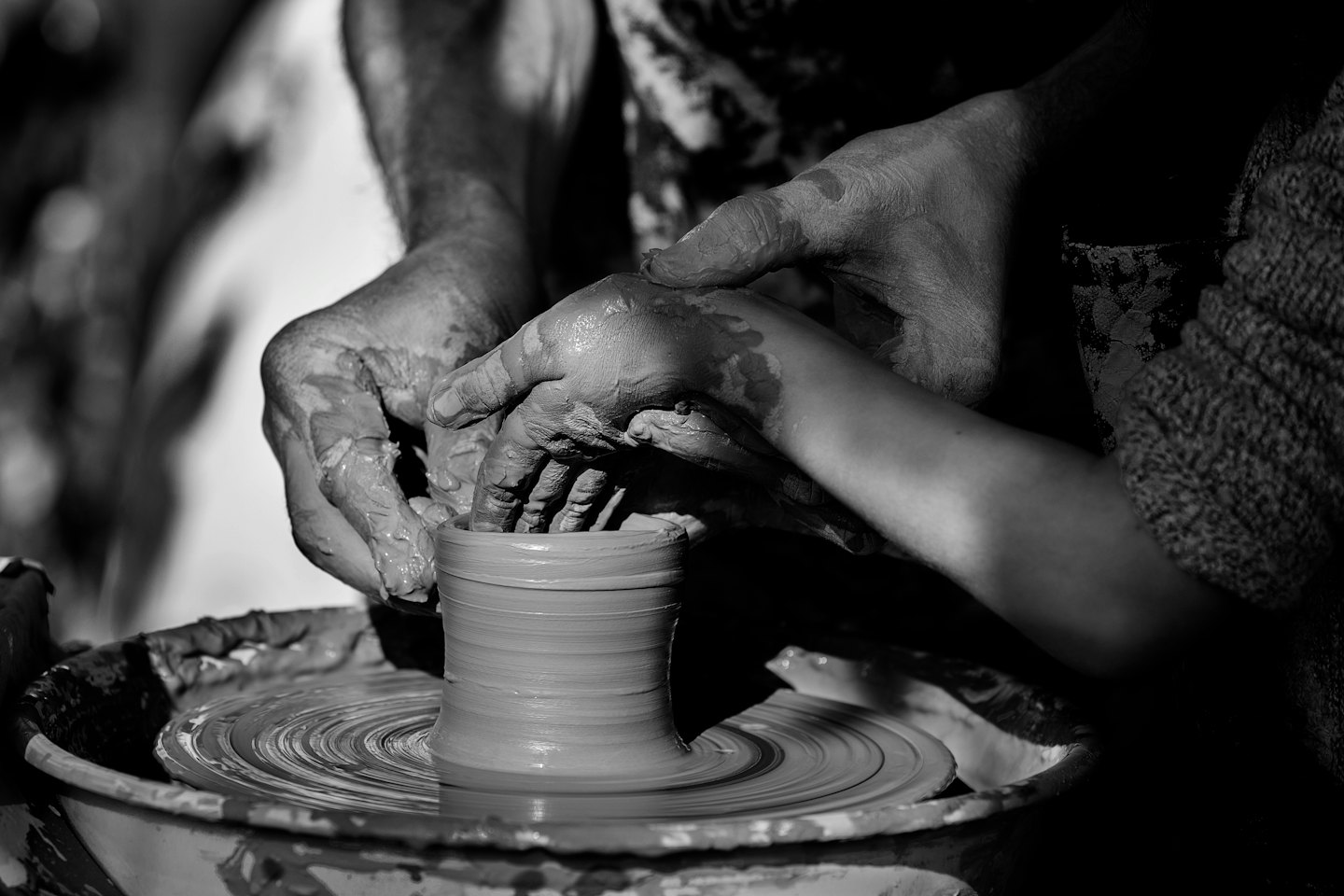 Adult and child's clay-covered hands shaping pottery together.